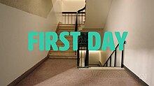 An long shot of an empty stairwell at a school. The words "First Day" are in the foreground in teal lettering.