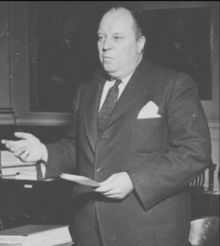 Perrow standing at desk motioning with right hand and holding paper in left hand