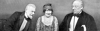 Three middle aged people in 1920s clothes, a woman centre, a man on each side, leaning towards her