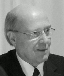 Three-quarter profile of a clean-shaven man with spectacles and receding hair, in suit and tie, in the act of talking.