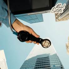 A photograph from below of a person's hand holding out a phone booth telephone with tall buildings in the background