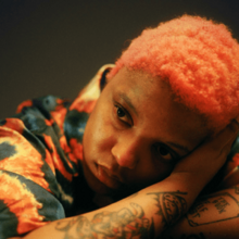 A photograph of James with short pink hair resting her head on her tattooed arm