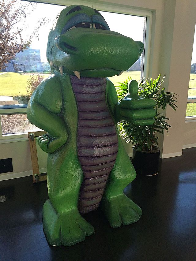 A photograph featuring the front right side of a statue of green lizard with a purple abdomen and chest. The lizard stands on two feet with its right arm on its hip and the left arm extended outward at the elbow, giving a thumbs up. Its eyes are partially closed and several sharp teeth extend from the closed mouth.