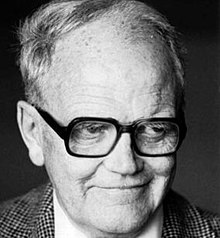 Black and white photo of a late middle-aged man with grey hair, wearing thick-rimmed eyeglasses, a white dress shirt, and a suitjacket in a checkered pattern