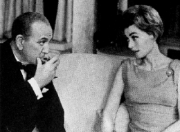 White man, balding, clean shaven, mid-sixties, in dinner jacket and black tie, sipping champagne alongside middle-aged white woman in evening dress