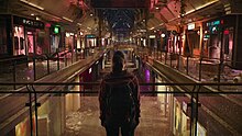 A girl with a backpack stands in the center of the frame as an abandoned mall lights up in front of her, with varying colors.