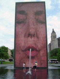 image of 50-foot-tall (15 m) architectural structure spouting water on children from a hole in its face. There is an image of a human face positioned such that the water appears to spout from the mouth. Blue sky and tall buildings are in the background.