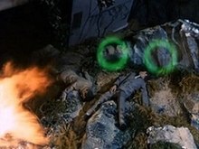 The broken bodies of two dead men lie on a rock face. Vehicle debris is strewn behind them and there is a burst of flame in the bottom-left corner. A pair of translucent green circles are passing over the men.