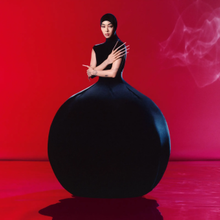 Rina Sawayama wears a black sculptural dress shaped like a ball in front of a red background