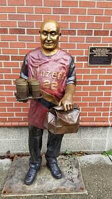 alt=Bronze statue of Todd Kirnan. He's depicted as holding a full 4-cup drink carrier in his right hand while his left hand grips the rolled top of a paper bag. He's dressed in a Portland Trailblazers basketball team jersey, #22. The statue is emplaced in front of a red brick wall. On the wall next to his left shoulder is a plaque that reads: "Mr. Gresham" Todd Kirnan Made possible with generous support of Heather and Rich Söderberg-Green Heart of the Gorge Bronze Söderberg Gallery and Studio The Citizens of Gresham Gresham Public Outdoor Art 2018