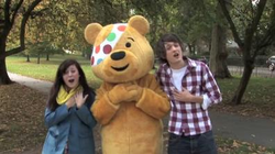 A young girl, a person dressed as a large smiling, yellow teddy bear and a young man stand in a grassy park, with their hands over their hearts. Their mouths are open, as if singing. The girl is wearing a blue jacket and a yellow scarf. The man is wearing a checked shirt and a white T-shirt. The teddy bear outfit has a polka dot bandana stitched over the bear's right eye.