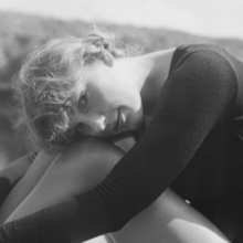A black-and-white-filtered picture of Swift looking towards the camera while hugging her knees