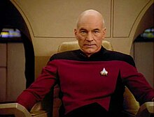 Tangkapan layar dari seorang pria botak ras putih berseragam militer fiksi ilmiah bernama Jean-Luc Picard; ia sedang duduk memandang kamera.