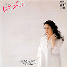 Một người phụ nữ Nhật Bản tóc đen, mặc áo sơ mi trắng, để tay lên vai cùng một phông nền tường màu trắng.