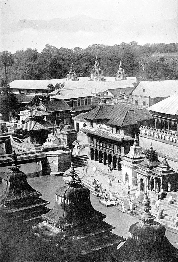 Black and white photograph, from a raised viewpoint, of a river lined by buildings. The bank of the river is paved with steps to the water itself. A foot bridge crosses the river at the left of the image. People are gathered below at the river bank. In the background, trees can be seen behind rooftops.