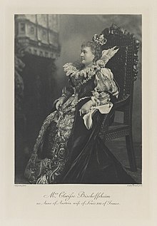 Black-and-white photograph of a seated woman richly dressed in an historical costume with a handkerchief in her left hand, a feather fan in her right and a crown on her head