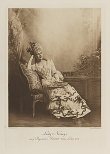 Black-and-white photograph of a seated woman richly dressed in an historical costume with her hair done in a tall pile of curls and a hat on top