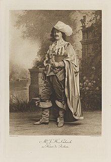 Black-and-white photograph of a standing man richly dressed in an historical costume with high boots, a sword, a long cloak, and a flamboyant hat