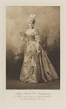 Black-and-white photograph of a standing woman richly dressed in an historical costume with long gloves, an open fan and feathers in her hair