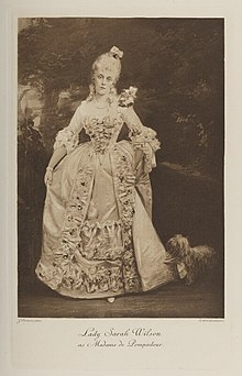 Black-and-white photograph of a standing woman richly dressed in an historical costume with a dog