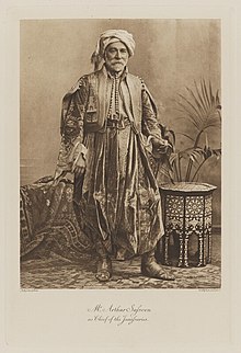 Black-and-white photograph of a standing man richly dressed in an historical, Middle-eastern costume