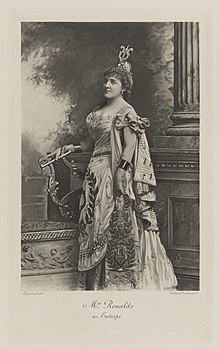 Black-and-white photograph of a standing woman richly dressed in a costume covered with symbols of music and carrying a lyre, with a lyre as part of her hat
