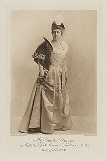 Black-and-white photograph of a standing woman richly dressed in an historical costume with a short cloak and a hat with a white feather plume