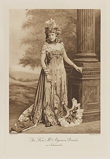 Black-and-white photograph of a standing woman richly dressed in an historical costume with a headdress and a very large fan