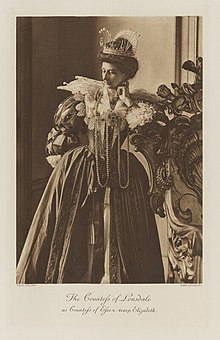 Black-and-white photograph of a standing woman richly dressed in an historical costume