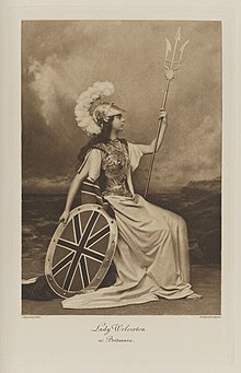 Black-and-white photograph of a seated woman dressed in an armor breast plate, with a shield, helmet and trident