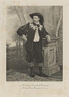 Black-and-white photograph of a standing man richly dressed in an historical costume with a wide-brim hat, a lacy collar and tie