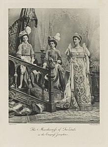 Black-and-white photograph of a standing woman richly dressed in an historical costume, with two boys dressed as pages