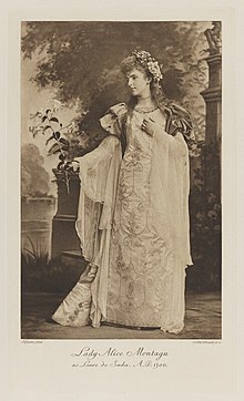 Black-and-white photograph of a standing woman richly dressed in an historical costume with flowers in her hair