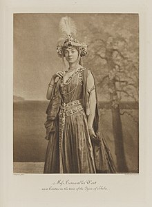 Black-and-white photograph of a standing woman richly dressed in an historical costume