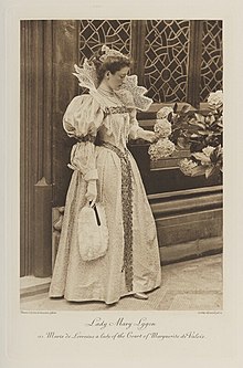 Black-and-white photograph of a standing woman richly dressed in an historical costume with a big, lacy color and a feather fan