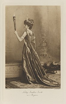 Black-and-white photograph of a standing woman with her back to the viewer, richly dressed in an historical costume holding a flaming torch, with crown on her head