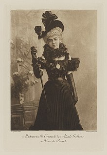 Black-and-white photograph of a standing woman richly dressed in a black historical costume
