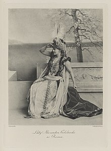 Black-and-white photograph of a seated woman richly dressed in an historical costume and shading her eyes to look into the distance