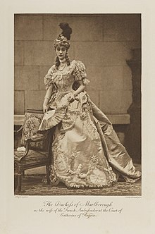 Black-and-white photograph of a standing woman richly dressed in an historical costume