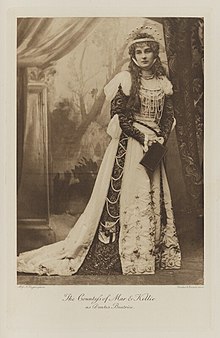 Black-and-white photograph of a standing woman richly dressed in an historical costume wearing a crown and holding a book
