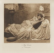 Black-and-white photograph of a reclining woman richly dressed in an historical costume and a crown on her head