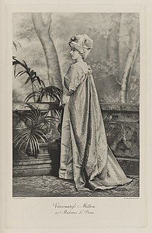 Black-and-white photograph of a standing woman richly dressed in an historical costume, mostly turned away and looking back over her left shoulder