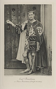 Black-and-white photograph of a standing man richly dressed in an historical costume, with armor, a long cape, and insignia of the Order of the Garter