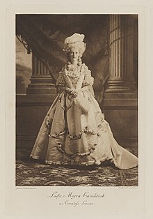 Black-and-white photograph of a standing woman richly dressed in an historical costume with a white wig and big hat