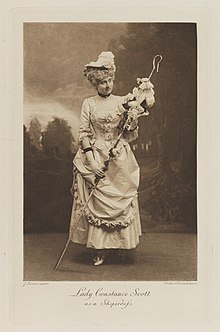 Black-and-white photograph of a standing woman richly dressed in an historical costume holding a staff with a hook at the end covered with flowers