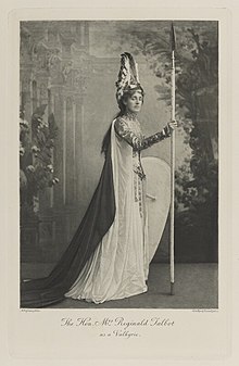 Black-and-white photograph of a standing woman richly dressed in an historical costume with a winged helmet, a shield and a long spear