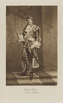 Black-and-white photograph of a seated man richly dressed in an historical military costume with knee-high boots and sword