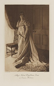 Black-and-white photograph of a standing woman richly dressed in an historical costume with a long train and a crown