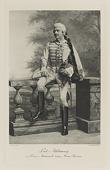 Black-and-white photograph of a man richly dressed in an historical military costume seated on a balustrade