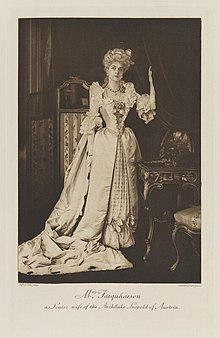 Black-and-white photograph of a standing woman richly dressed in an historical costume with a long train